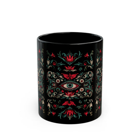 A black ceramic mug featuring a symmetrical folk art design with a central mystic eye motif in turquoise and coral. The design includes ornate scrollwork, red tulip-like flowers, sage green leaves, and cream flourishes arranged in a decorative pattern. The mug has a glossy finish with a C-shaped handle.
